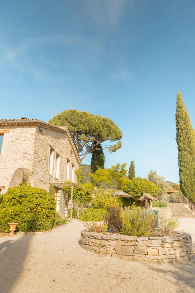 Jardin méditerranéen aménagé devant un mas en pierre avec pin parasol, cyprès élancé et murets en pierres sèches sous un ciel bleu.
